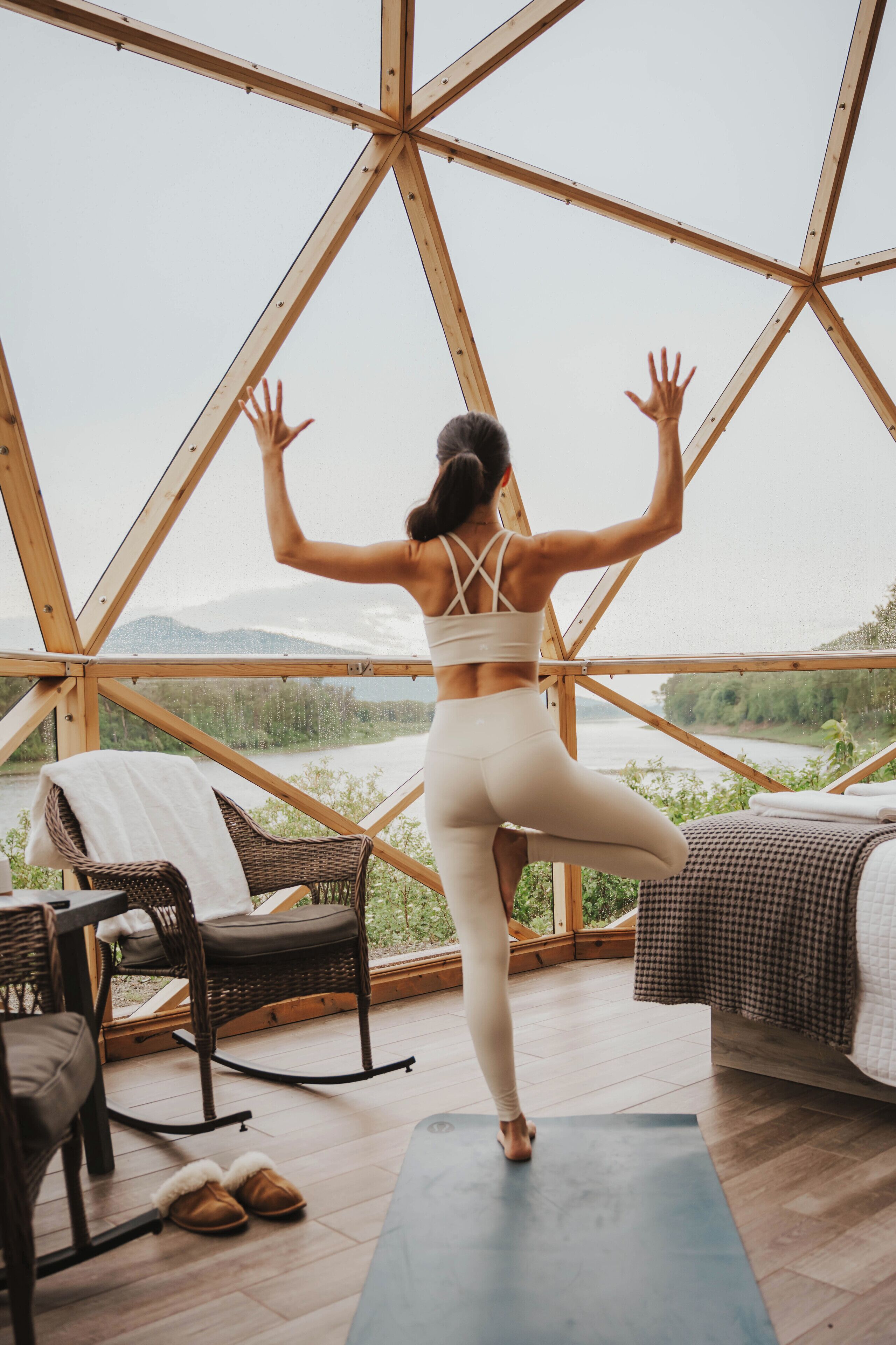 A woman performing a yoga pose on a mat inside a geodesic dome structure with a view of a river and mountains.