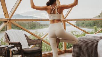 A woman performing a yoga pose on a mat inside a geodesic dome structure with a view of a river and mountains.