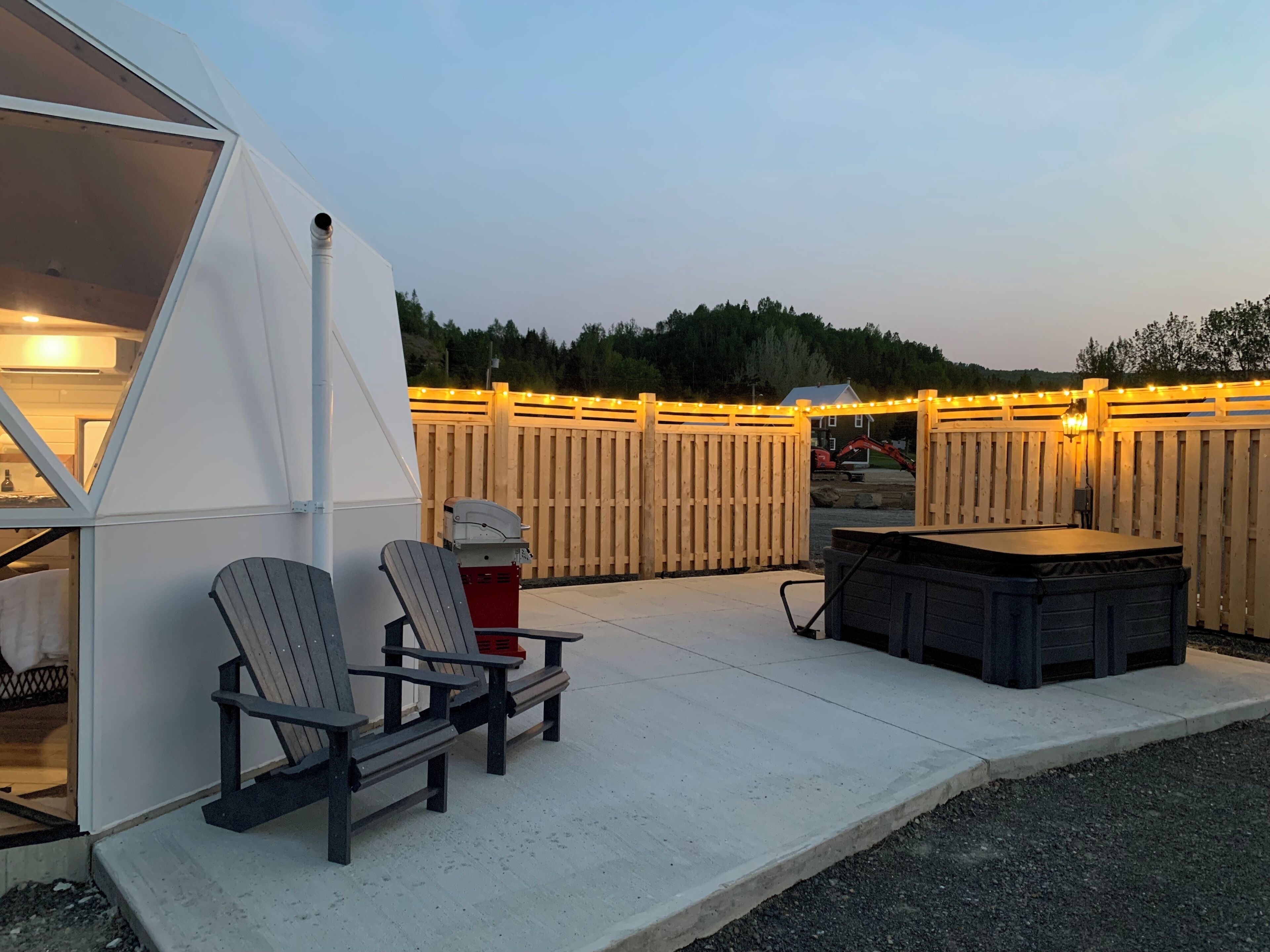 Un espacio al aire libre cubierto con sillas tipo Adirondack, un jacuzzi y una valla de madera con luces colgantes.
