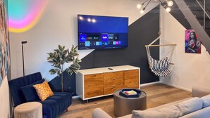 Egy modern nappali, melyben található egy síkképernyős televízió, egy függő szék, egy kanapé, egy dohányasztal és egy lépcső.