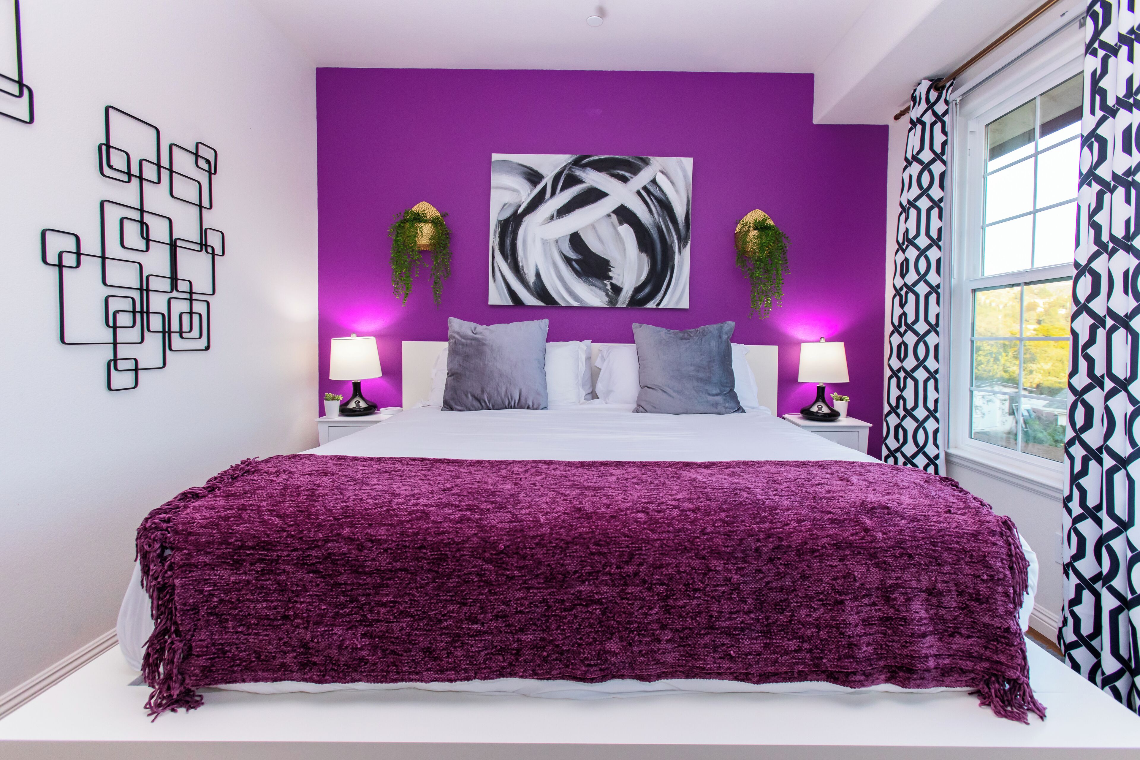 A bedroom with a purple accent wall, a large abstract painting, two bedside lamps, and a window with patterned curtains.