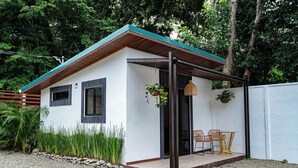 A small, single-story house with a white exterior and a green roof. It features a porch with wicker chairs and a hanging plant. - Villas Dìwö (Cahuita)