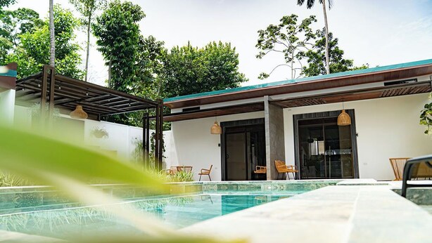 A swimming pool with a pergola and chairs, surrounded by lush greenery and a modern building with large windows. - Villas Dìwö (Cahuita)