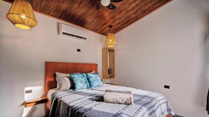 A bedroom with a wooden ceiling, a bed with a striped bedspread, two bedside lamps, and a nightstand. - Villas Dìwö (Cahuita)