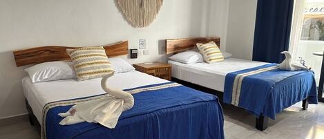 A hotel room with two beds, each with a blue bedspread and a white swan-shaped towel. There is a macrame wall hanging above the beds and a wooden headboard.