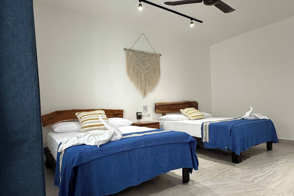 Une chambre d’hôtel avec deux lits, chacun recouvert d’une couverture bleue et agrémenté d’oreillers blancs. Un tableau est accroché au mur au-dessus des lits, et un ventilateur de plafond est installé.