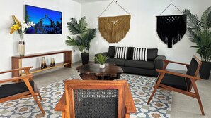 Ein modernes Wohnzimmer mit einer Couch, einem Couchtisch und Sesseln. Es gibt einen Deckenventilator, einen an der Wand montierten Fernseher und Zierpflanzen.