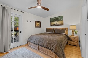 A bedroom with a bed, a ceiling fan, a nightstand with a lamp, a painting on the wall, and a door with a view of the outdoors.