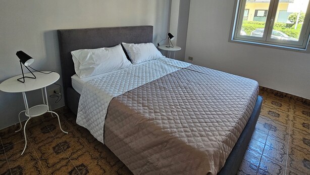A neatly made bed with a quilted cover and pillows, a small bedside table with a lamp, and a window with a view of buildings and greenery. - Guests' Nest (Castellammare del Golfo)