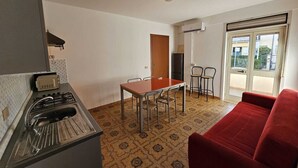 A compact kitchen with a gas stove and sink, a dining table with chairs, a red sofa, and a sliding glass door leading to a balcony. - Guests' Nest (Castellammare del Golfo)