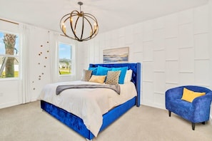 A bedroom with a large bed, a blue armchair, a chandelier, and a view of the outdoors. - 19 Guest Home w Pool Hot Tub Cinema BBQ  (Kissimmee)