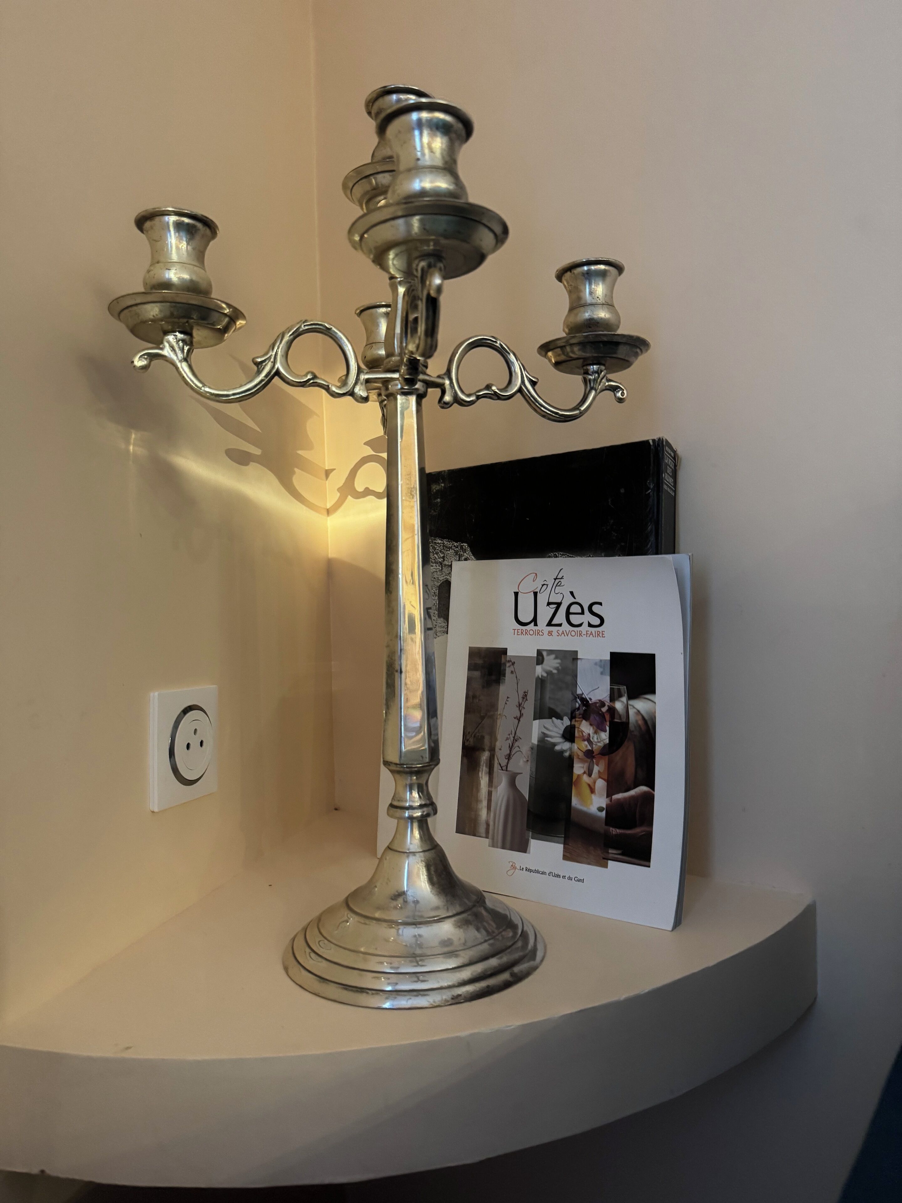 A silver candelabra with four candles on a white shelf, next to a book titled 'Uzes'.