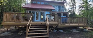A two-story wooden cabin with a red roof, a deck, and a staircase leading to the entrance. - 4 Season Lakefront Cottage with Hot Tub (Little Deer Lake)