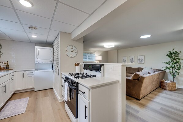 A modern kitchen with white cabinets, a stove, and a refrigerator. There is a dining area with a brown sofa and a plant.
