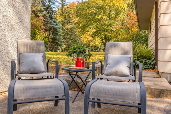 Two outdoor lounge chairs with cushions, a small round table, and a bottle of wine, set against a backdrop of trees and a house.