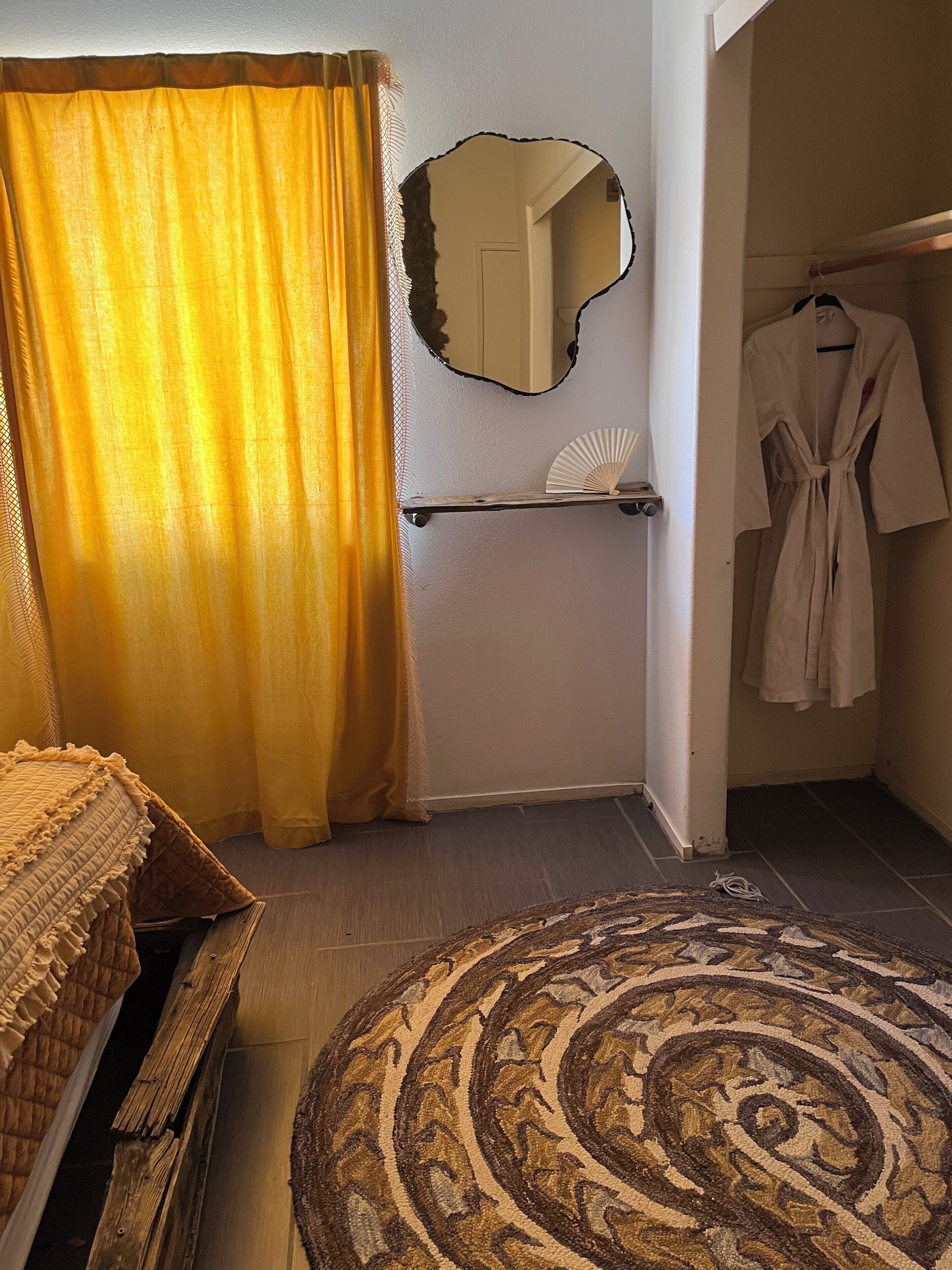 A bedroom with a bed, a round patterned rug, a mirror, a robe hanging, and a shelf with a fan.