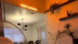 A living room with a hanging chair, a dining area with chairs, a wall-mounted shelf with plants, and a neon sign reading "DESERT DIMENSION".