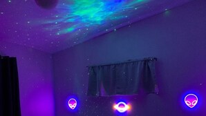 A bedroom with a bed, two bedside tables, a window with curtains, and a ceiling projection of stars and aurora.