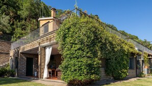 Un bâtiment en pierre doté d’un espace extérieur abrité avec des sièges, d’un escalier et d’un grand arbuste à l’avant.