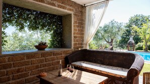 Un espace extérieur accueillant avec un canapé en osier et une table en bois, un mur en briques, une plante en pot et une vue sur une piscine et des arbres.