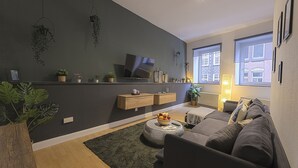 Una sala de estar moderna con pared oscura, piso de madera y un sofá gris. Hay un televisor en la pared, una mesa de centro redonda con fruta y una mesita auxiliar.