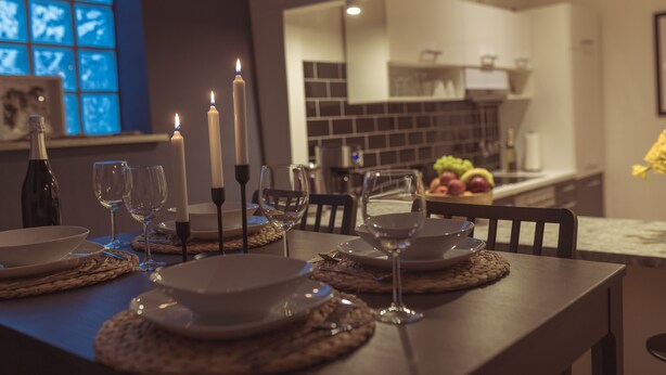 Una mesa de comedor dispuesta con platos, vasos y velas, con una cocina a la vista al fondo.