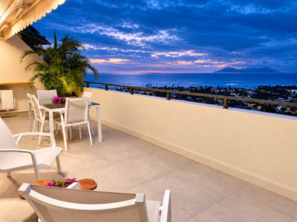 A balcony with a table and chairs, overlooking a coastal cityscape at dusk.