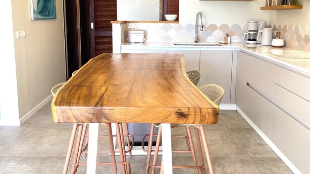 A modern kitchen with a wooden dining table, copper bar stools, pendant lights, and a white countertop.