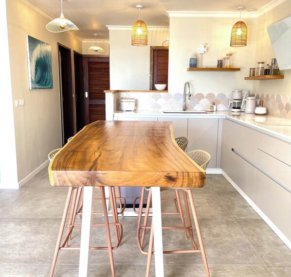 A modern kitchen with a wooden dining table, copper bar stools, pendant lights, and a white countertop.