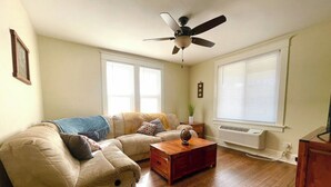 Ein Wohnzimmer mit einem Ecksofa, einem Holztisch, einem Deckenventilator und einem großen Fenster mit Jalousien.