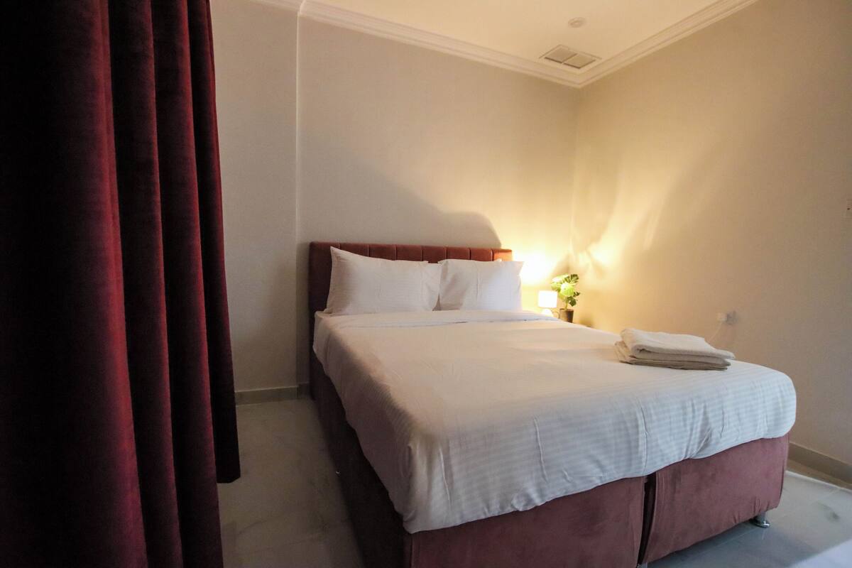 A hotel room with a bed, curtains, a nightstand with a lamp, and a folded towel on the bed.
