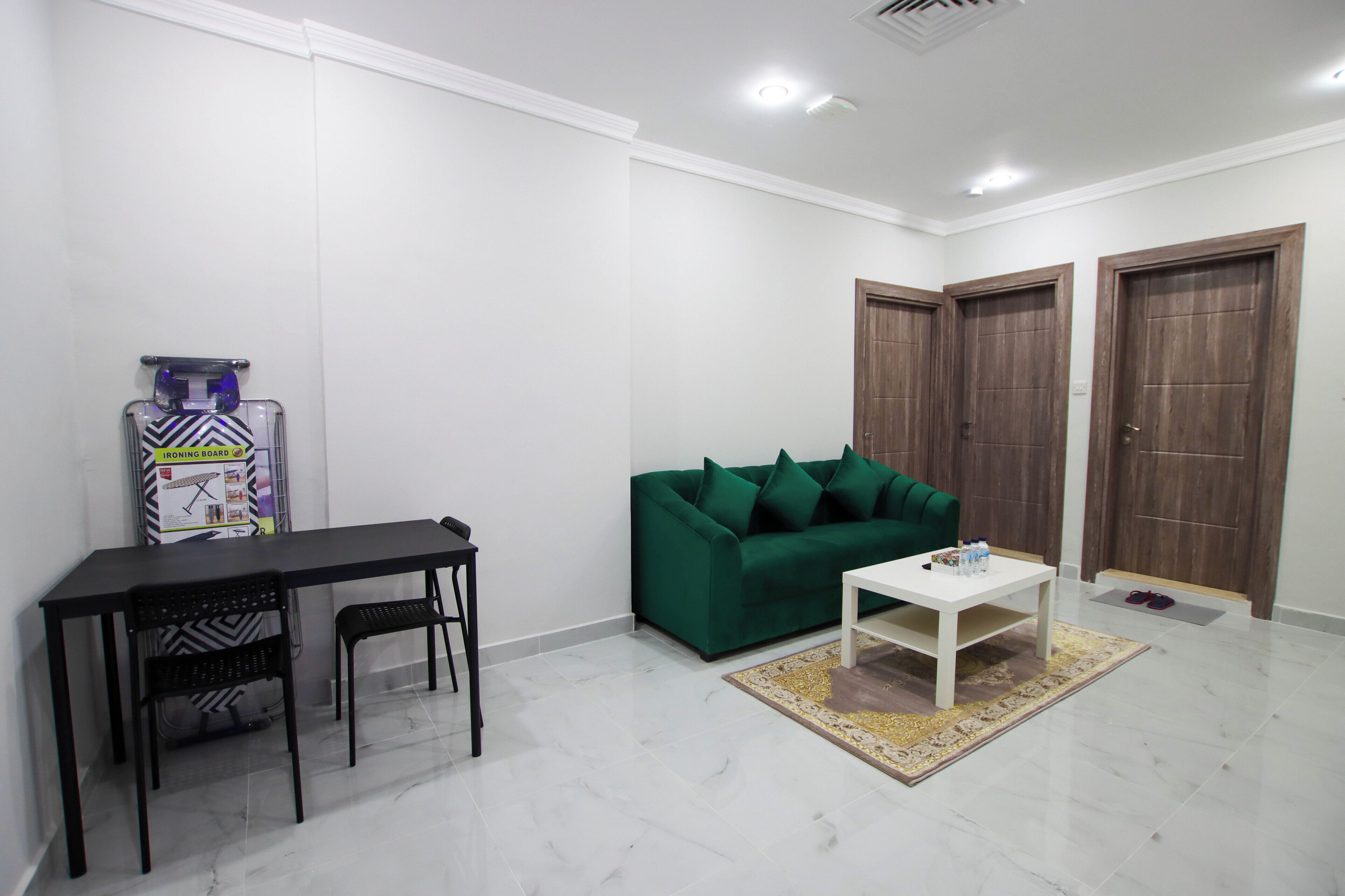 Sala de estar com sofá verde, mesa de centro branca, mesa de jantar preta e uma cadeira preta. Ao fundo, uma porta de madeira e uma parede branca.