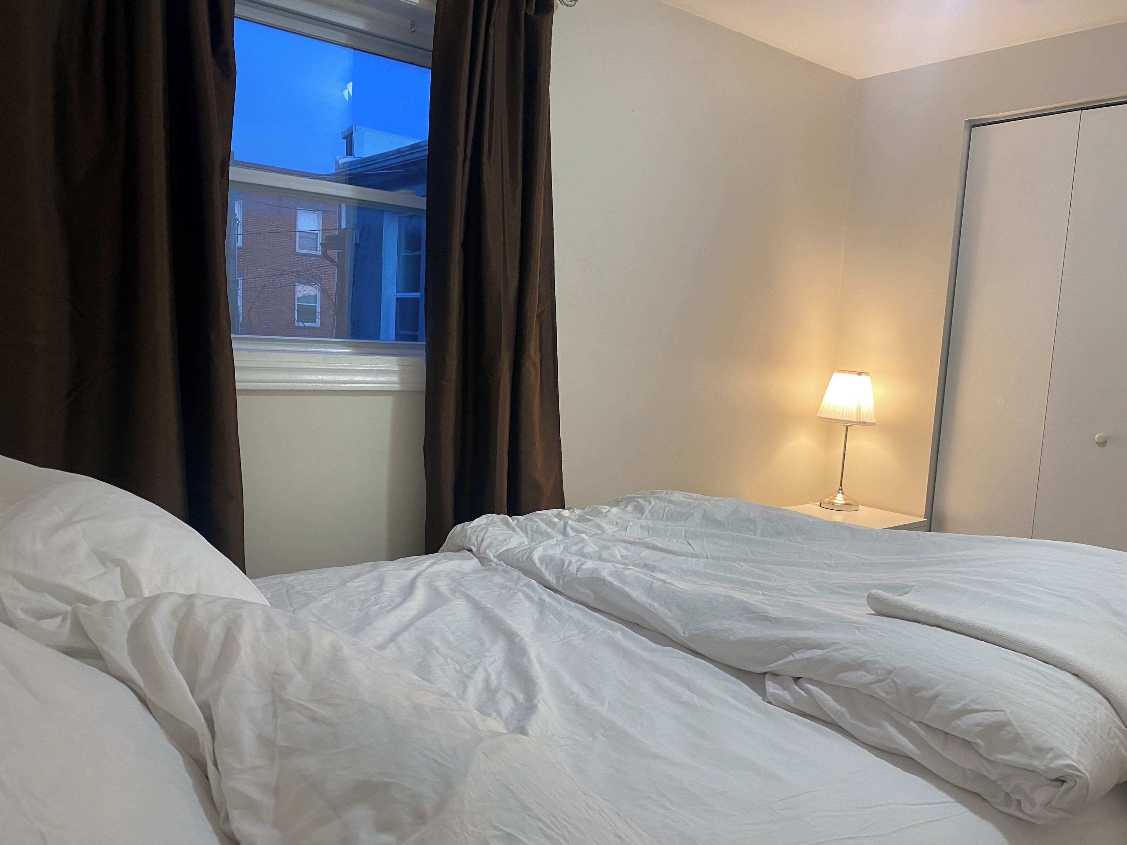 A hotel room with a bed, a window with curtains, a nightstand with a lamp, and a wardrobe.