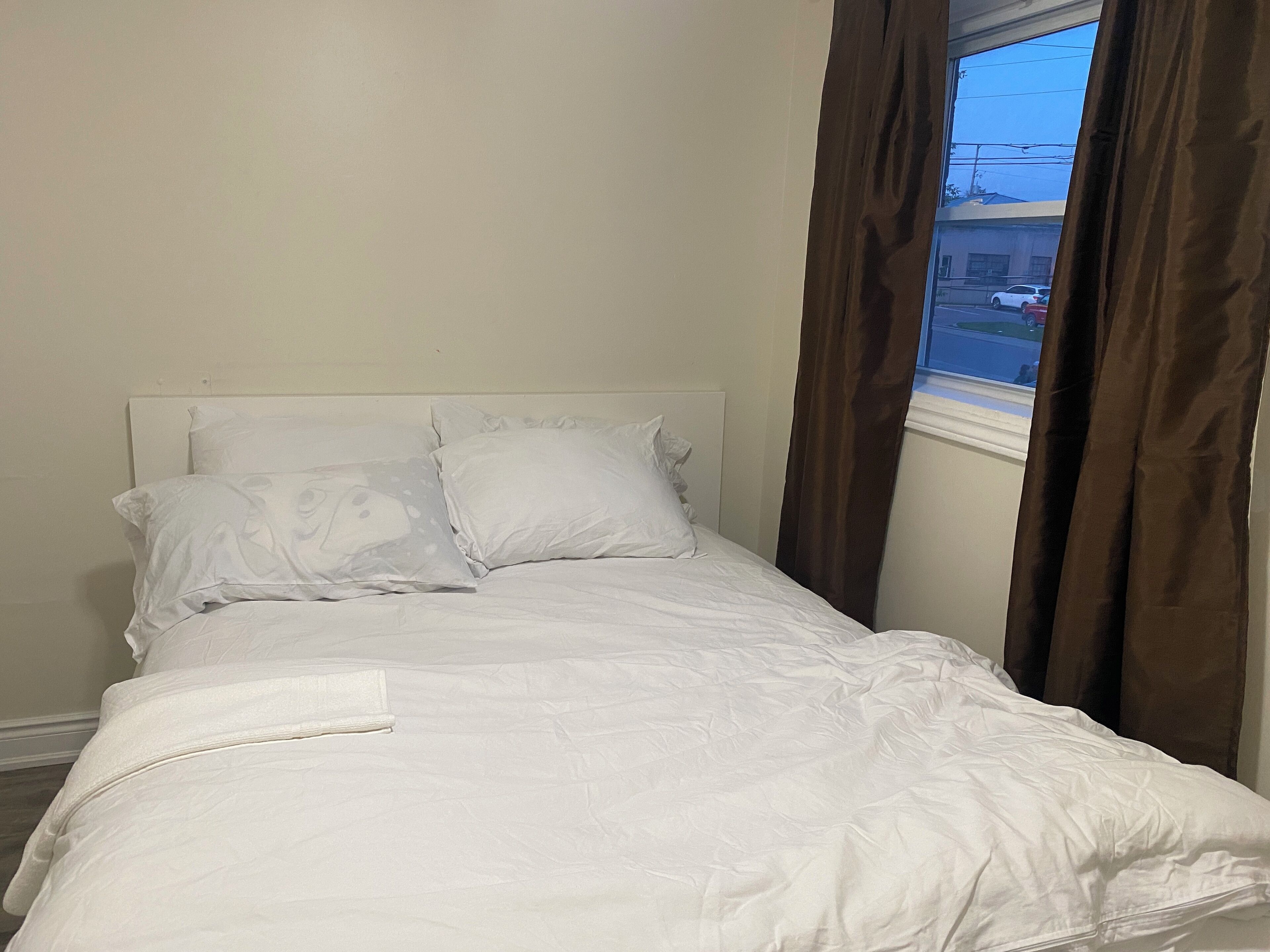 Een netjes opgemaakt bed met witte lakens en twee kussens, een raam met gordijnen en een effen muur.