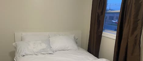 Een netjes opgemaakt bed met witte lakens en twee kussens, een raam met gordijnen en een effen muur.