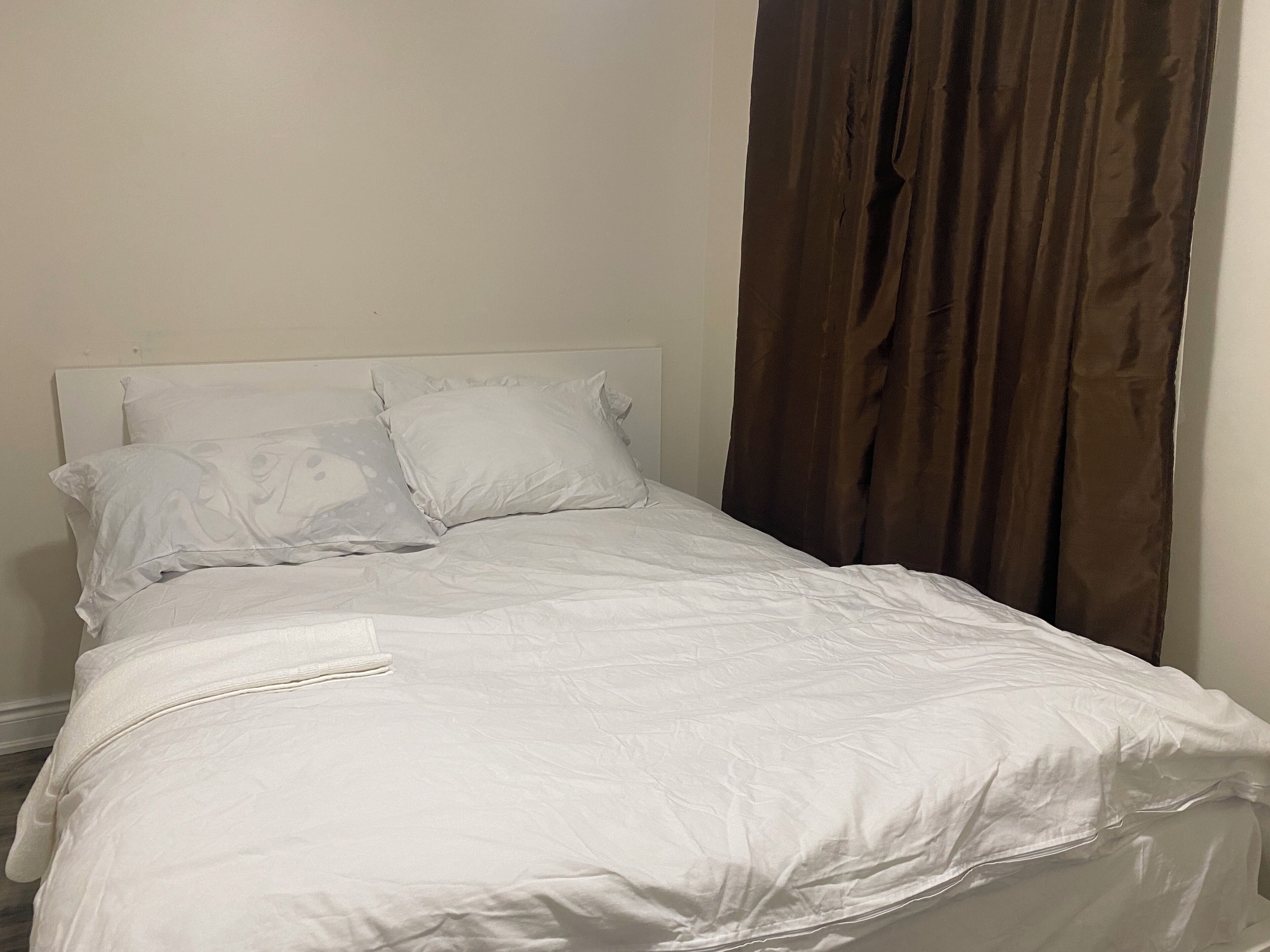 Una cama bien hecha con sábanas y almohadas blancas, un cabecero sencillo y una cortina marrón a la derecha.