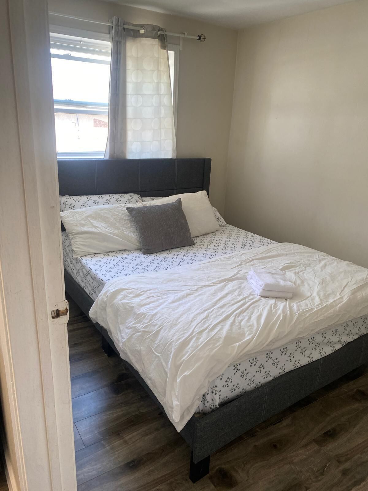 Un letto rifatto con lenzuola bianche e un piumone a motivi floreali, una testiera scura e una finestra con tenda.