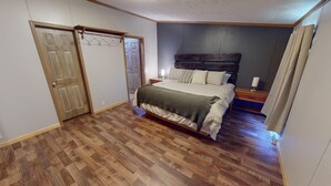 A bedroom with a wooden floor, a bed with a green blanket, a nightstand with a lamp, and a wooden headboard. - “Paradise Hill”  A Charming 3-bedroom house in the country. (Victor)