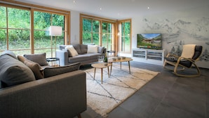 Ein modernes Wohnzimmer mit grauem Sofa, einem Holztisch und einem großen Fenster mit Blick auf Grünflächen.