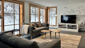 Ein modernes Wohnzimmer mit einem grauen Sofa, einem Couchtisch und einem Fernseher auf einer weißen Kommode. Große Fenster bieten Blick auf eine verschneite Landschaft.