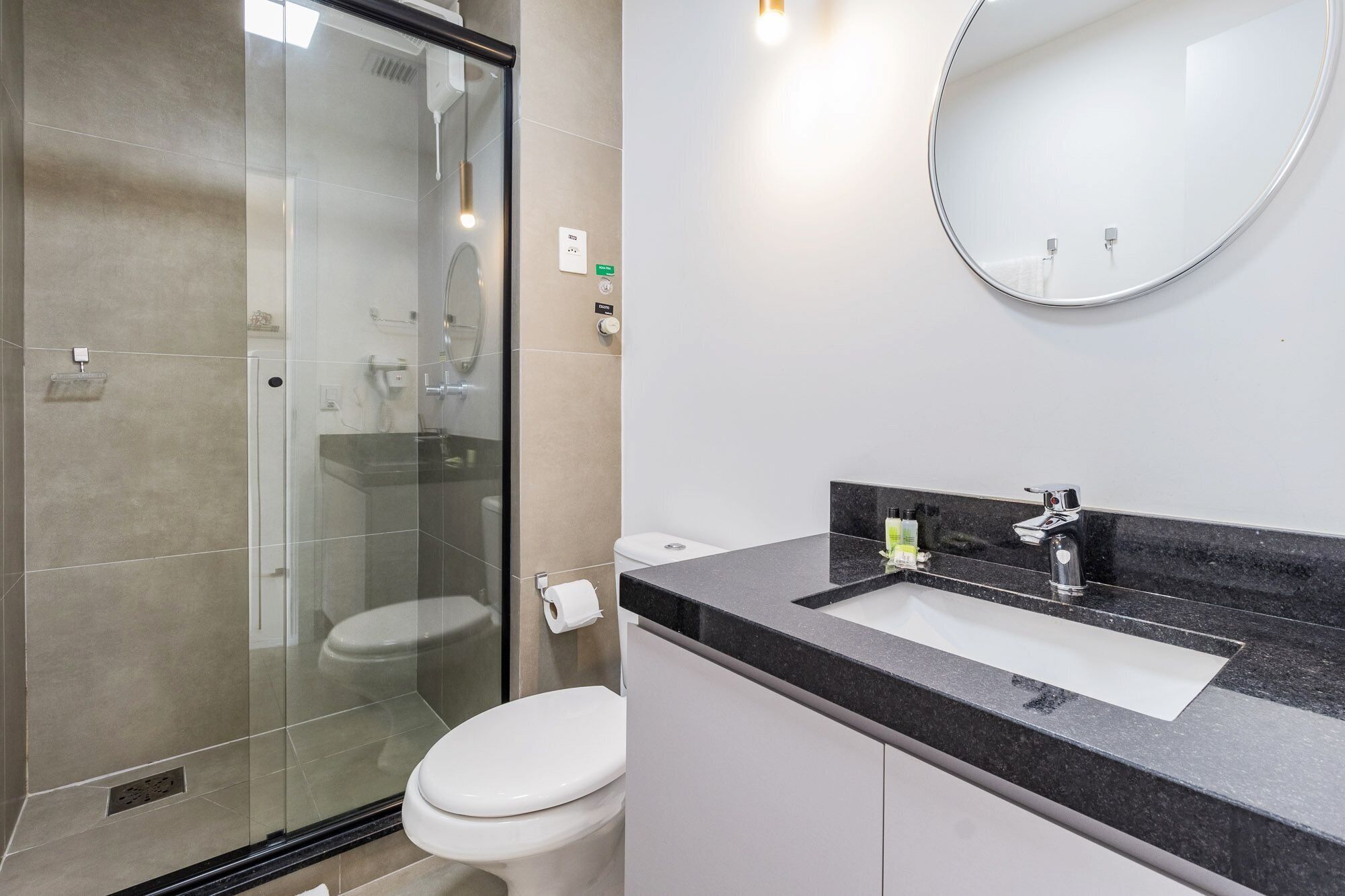 A modern bathroom with a glass shower enclosure, a white toilet, a sink with a black countertop, and a round mirror.