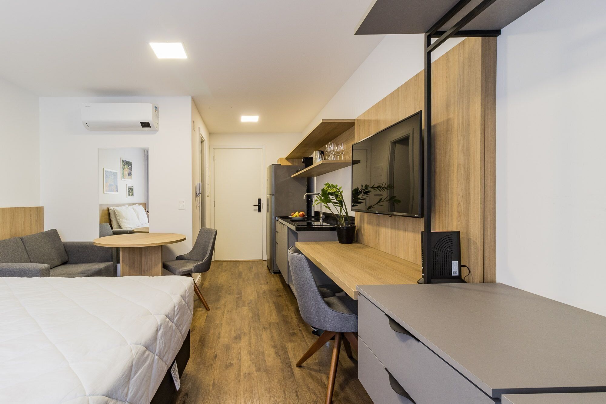 Quarto de hotel compacto com cama, escrivaninha, cadeira, mesinha, televisão e cozinha compacta.