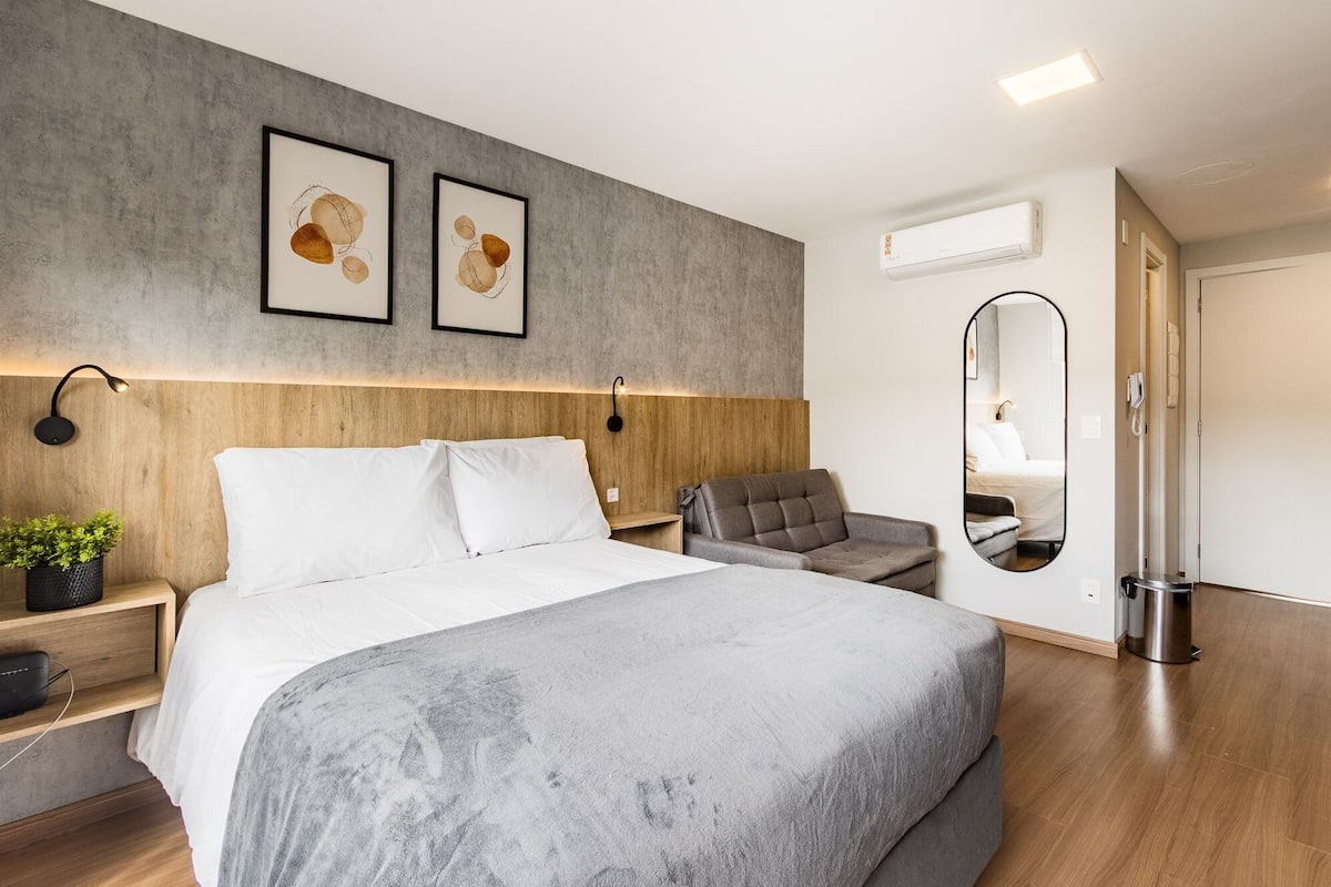 Una habitación de hotel moderna con una cama grande, un sofá gris, una mesita con una planta, un espejo en la pared y una lámpara en el techo.