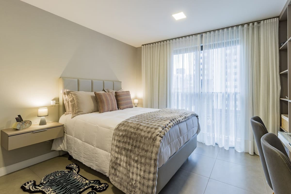 Habitación de hotel con cama, lámparas de noche, mesita de noche, una silla y vistas a la ciudad a través de cortinas translúcidas.