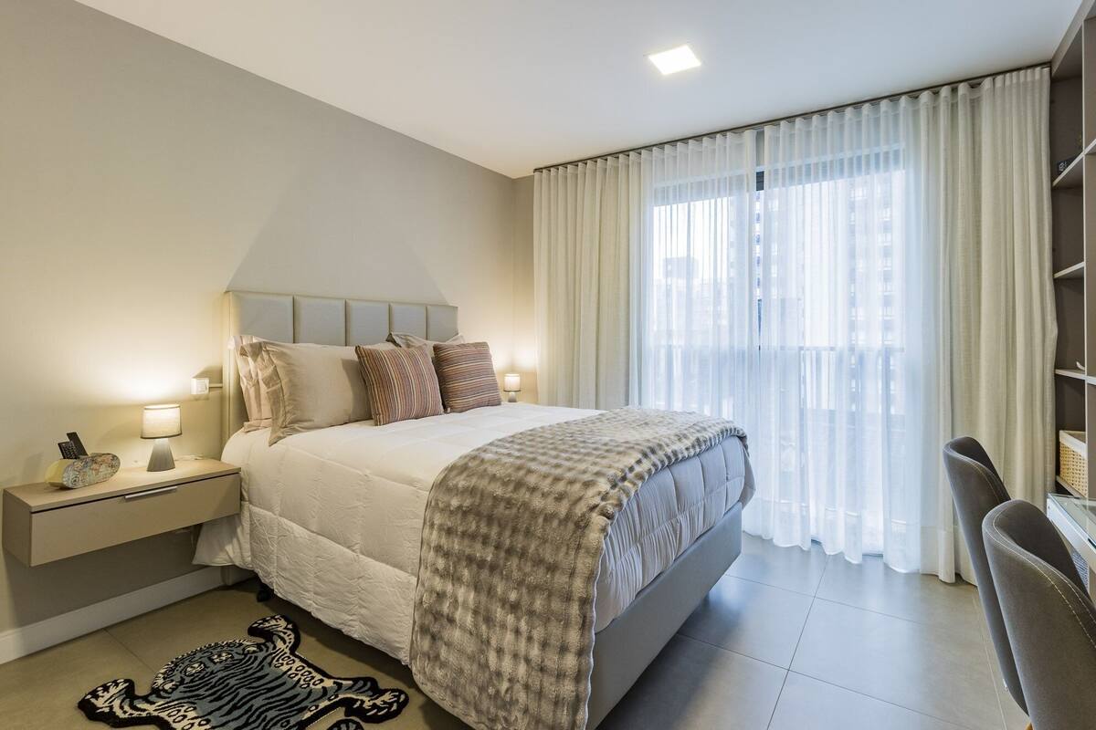 Camera d'albergo con un letto, lampade da comodino, un tavolino, una sedia e vista sulla città attraverso tende leggere.