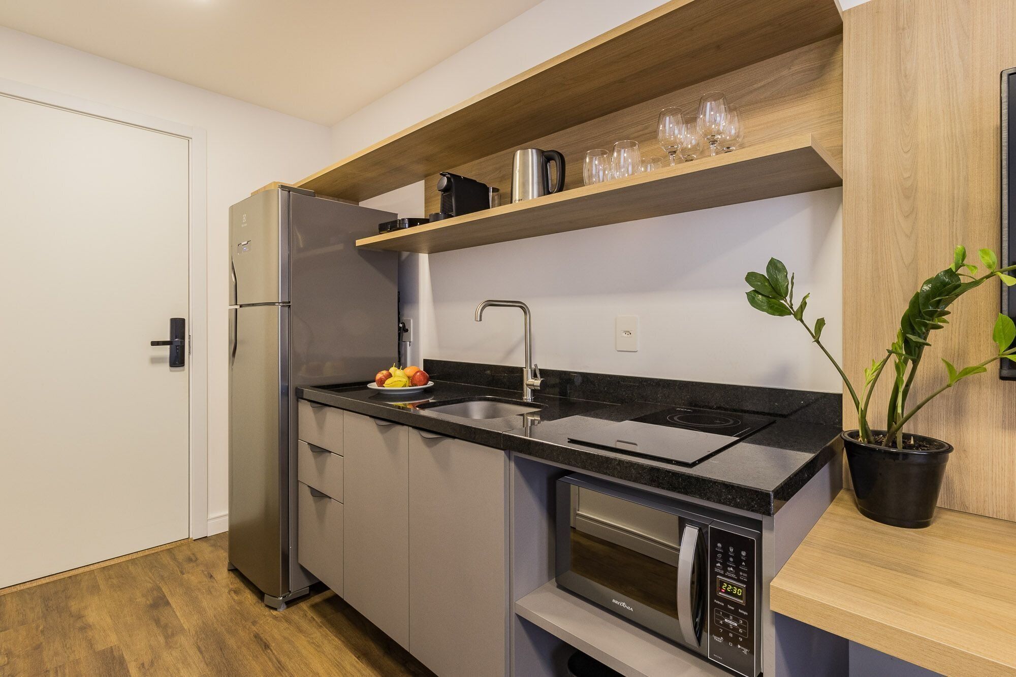 Eine kleine Küche mit Kühlschrank, Backofen, Spüle sowie einem Regal mit Gläsern und Wasserkocher.