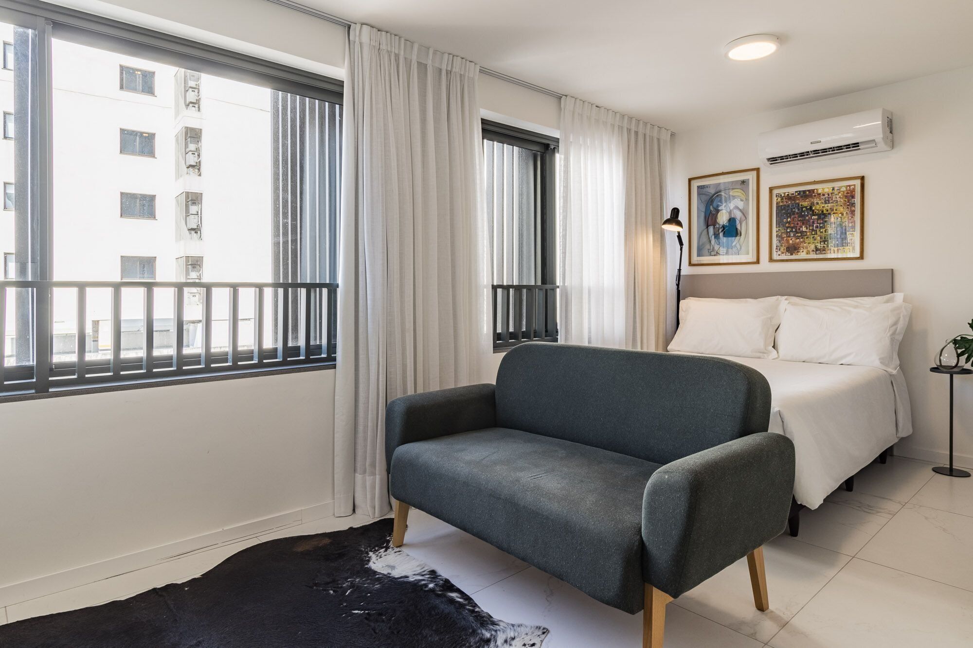 A hotel room with a sofa, a bed, a black and white rug, a window with curtains, and two framed pictures on the wall.