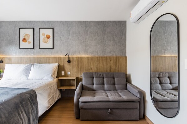 Una habitación de hotel moderna con una cama, un sillón gris, una pequeña mesita de noche y un espejo grande.