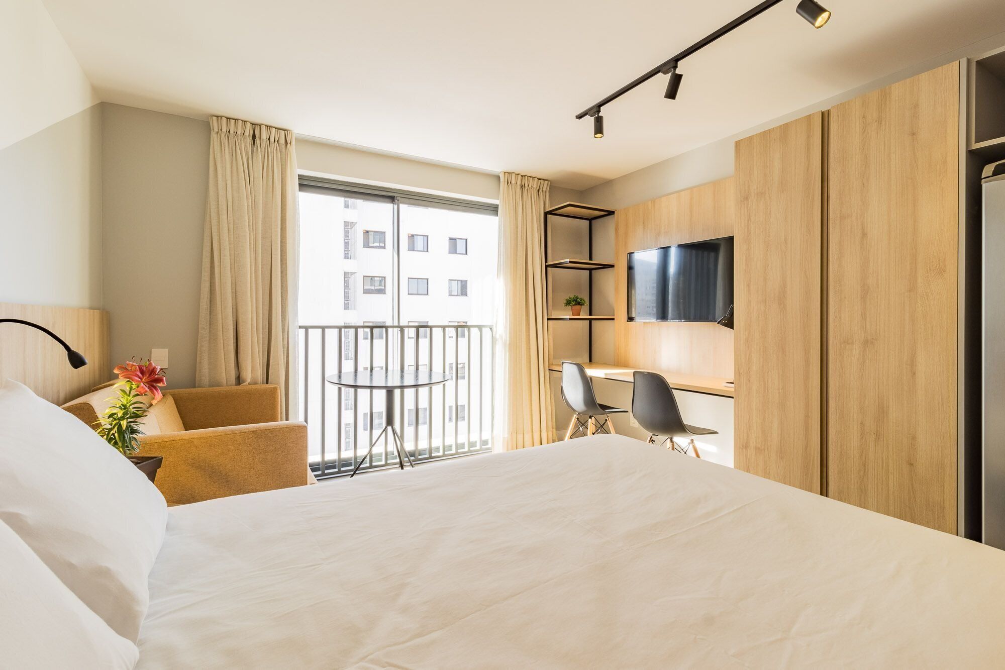 Ein Hotelzimmer mit einem Bett, einem Schreibtisch mit zwei Stühlen, einem Fernseher und einem Balkon mit Vorhängen.