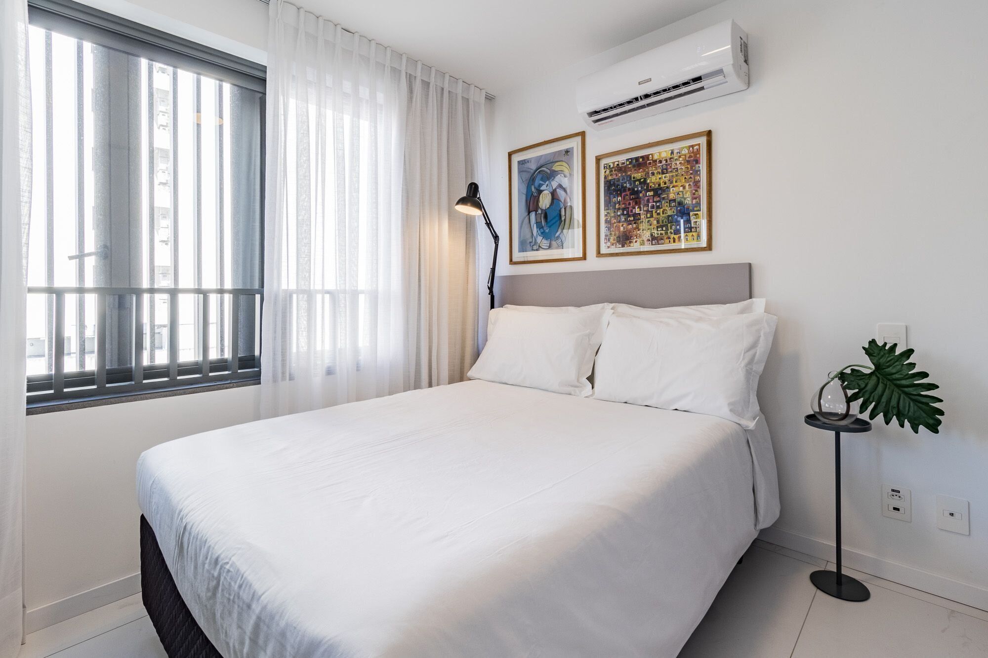 Ein Hotelzimmer mit einem Bett, einem Fenster mit Jalousien, einer Klimaanlage, zwei gerahmten Kunstwerken, einer Stehlampe und einer Topfpflanze.